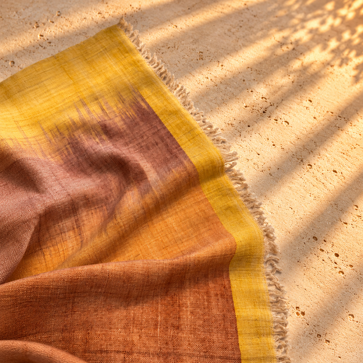 A close-up of a handwoven pashmina ikat foulard with a warm gradient from mustard yellow to terracotta and dusty rose, draped on a sandy surface with palm leaf shadows.