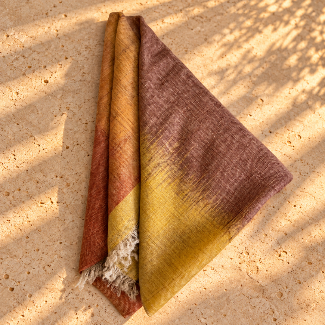 A folded woven Pure pashmina scarf foulard with a warm gradient from terracotta and mustard yellow to dusty mauve, laid on a sunlit stone surface with dappled shadows.