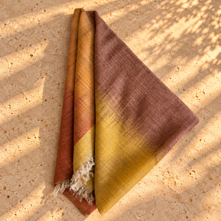 A folded woven Pure pashmina scarf foulard with a warm gradient from terracotta and mustard yellow to dusty mauve, laid on a sunlit stone surface with dappled shadows.