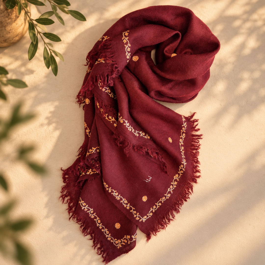 A deep maroon Pashmina foulard with gold embroidered borders and motifs, styled in a draped flat-lay on a stone surface with soft leaf shadows.