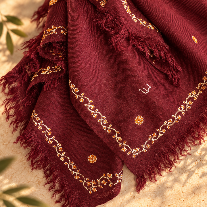 A deep burgundy scarf with gold floral embroidery and fringed edges, draped on a sandy surface with soft green leaves in the background.