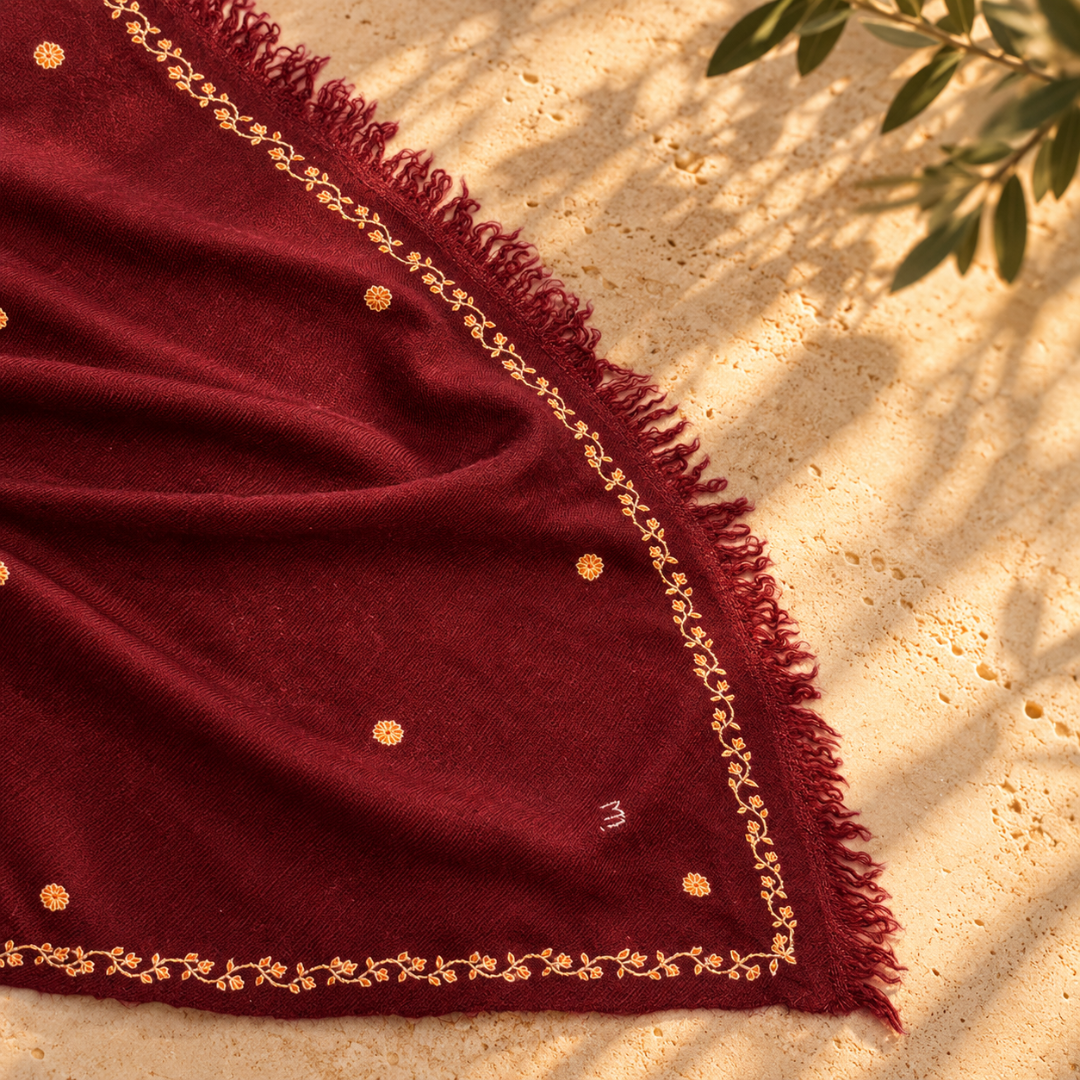 A deep burgundy Pashmina foulard scarf with gold embroidered borders and small gold dots, draped on a sandy surface with leaf shadows.