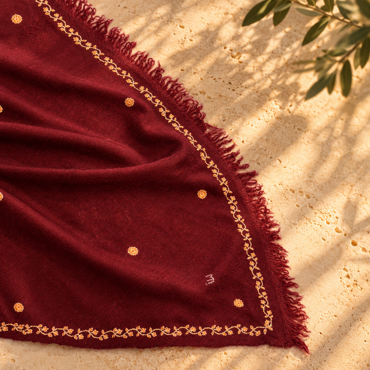 A deep burgundy Pashmina foulard scarf with gold embroidered borders and small gold dots, draped on a sandy surface with leaf shadows.