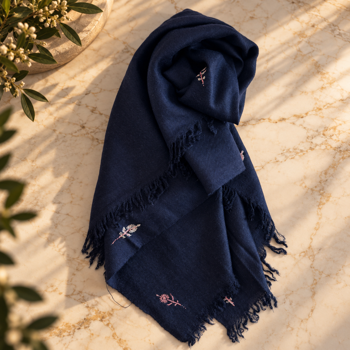 A navy blue fringed scarf with delicate embroidered floral details, styled on a beige marble surface with soft leaf shadows.