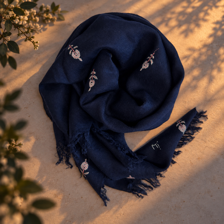 A dark navy blue scarf with small white pink embroidered motifs and fringed edges, styled on a stone surface with warm sunlight and leaf shadows.
