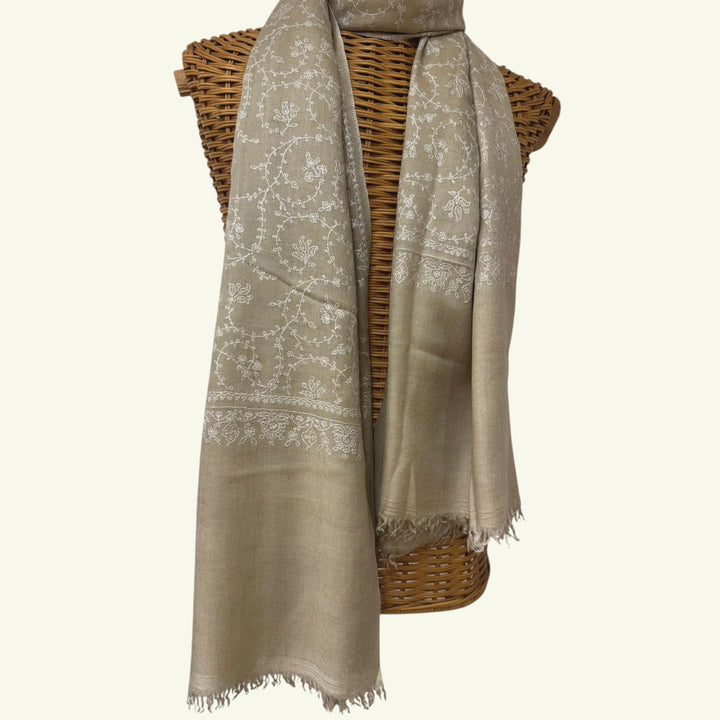 A light beige scarf with white floral embroidery and fringed edges, draped over a brown woven wicker stand against a plain off-white background.