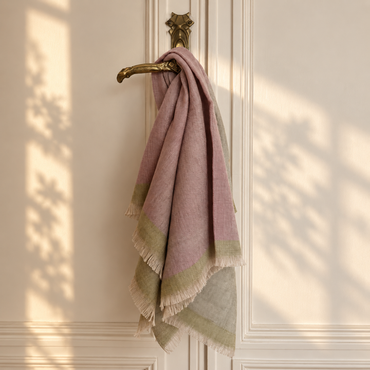 A soft handwoven pashmina foulard scarf  in muted dusty pink and sage green hangs from a brass door handle against an off-white paneled door, with dappled sunlight and leaf shadows cast across the surface.