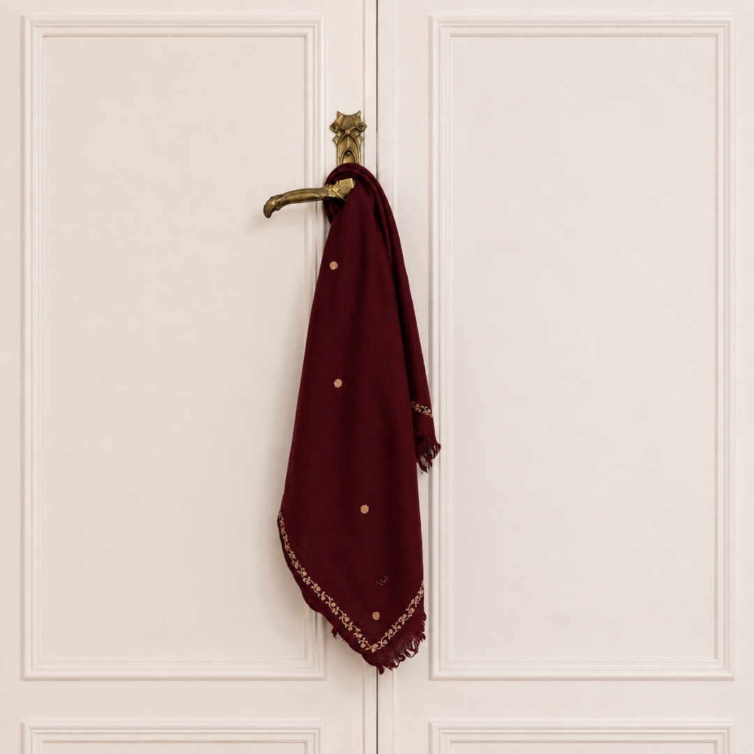 A deep maroon scarf with gold embroidered details and small gold floral dots, hanging from a brass doorknob on a white paneled door.