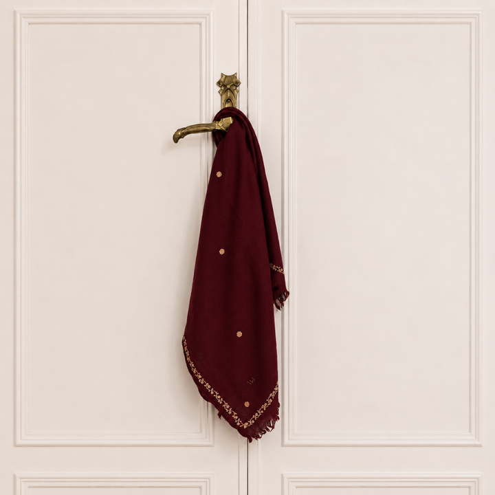A deep maroon scarf with gold embroidered details and small gold floral dots, hanging from a brass doorknob on a white paneled door.