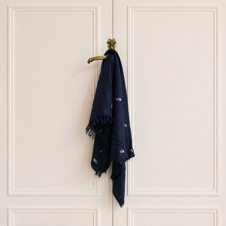 A navy blue scarf with small white embroidered motifs and fringed edges hangs from a brass door hook on cream-colored paneled doors.