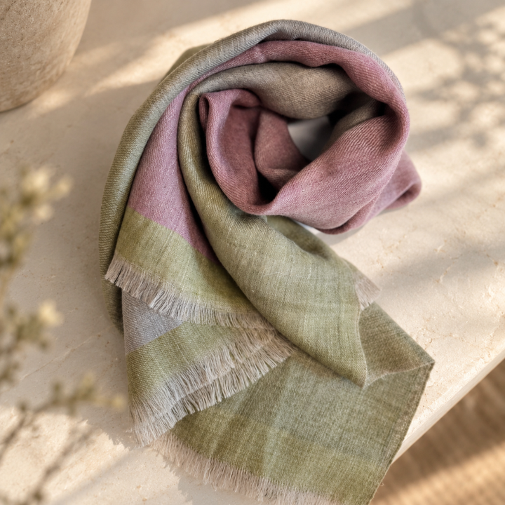 A folded handwoven indusweavers scarf with fringed edges in soft muted tones of dusty pink, sage green, and light gray, styled on a light stone surface with soft natural shadows.