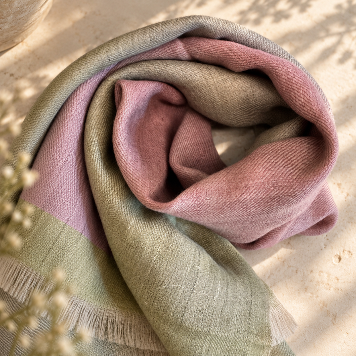 A soft pashmina handwoven foulard draped in a loose spiral, showing a ikat color transition of dusty pink, sage green, and light beige. The scarf has a fringed edge and is styled on a textured beige stone surface with soft shadows and dried flowers in the foreground.
