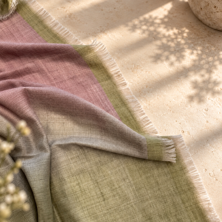 A soft woven pashmina scarf foulard with fringed edges draped on a light stone surface, showing a color-blocked design in dusty pink, sage green, and light gray-beige, with dappled sunlight and leaf shadows across the scene.