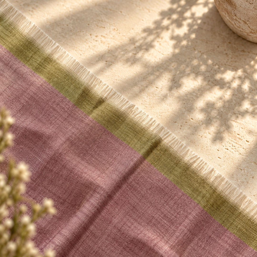 A close-up of pure Pashmina Foulard laid diagonally across a light stone surface, with a dusty mauve-pink main field, an olive-green border, and white fringe along one edge. Sunlight and leaf shadows fall across the scene, with small dried flowers in the foreground.