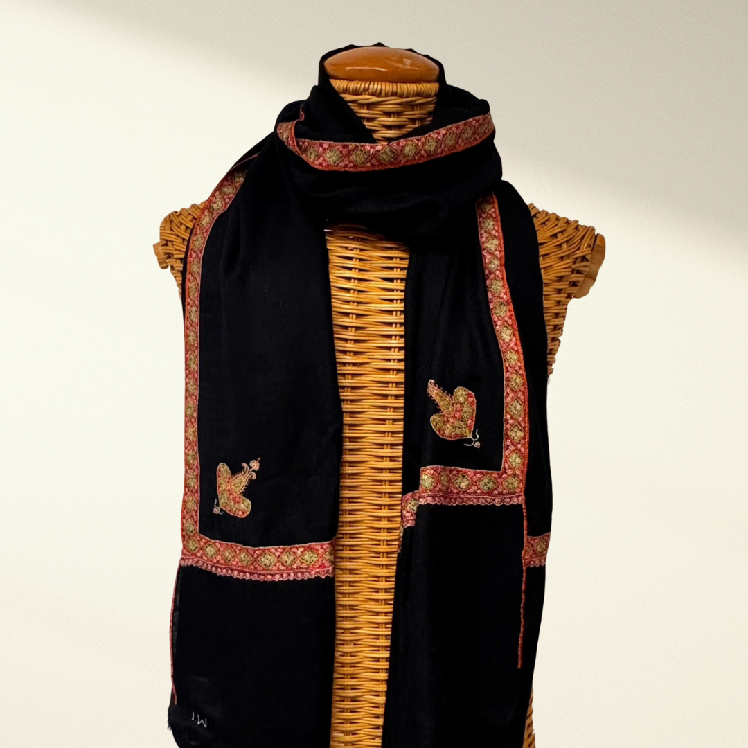 Black pashmina scarf with floral patterns and bird designs draped over a woven basket on a light background