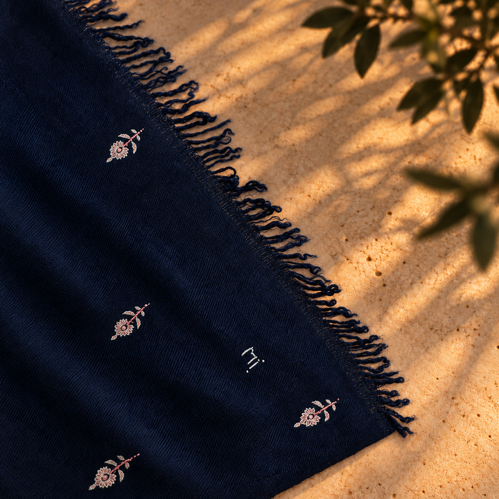 A close-up of a dark navy blue scarf with delicate embroidered floral motifs and a fringed edge, laid diagonally on a sandy surface with dappled sunlight and leaf shadows.