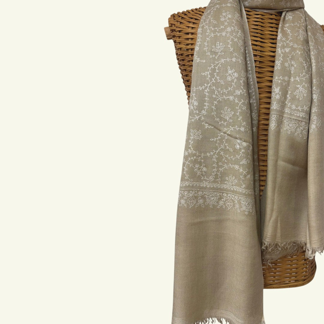 A light taupe-beige shawl with white floral embroidery and fringed edges, draped over a brown woven wicker basket against a plain off-white background.