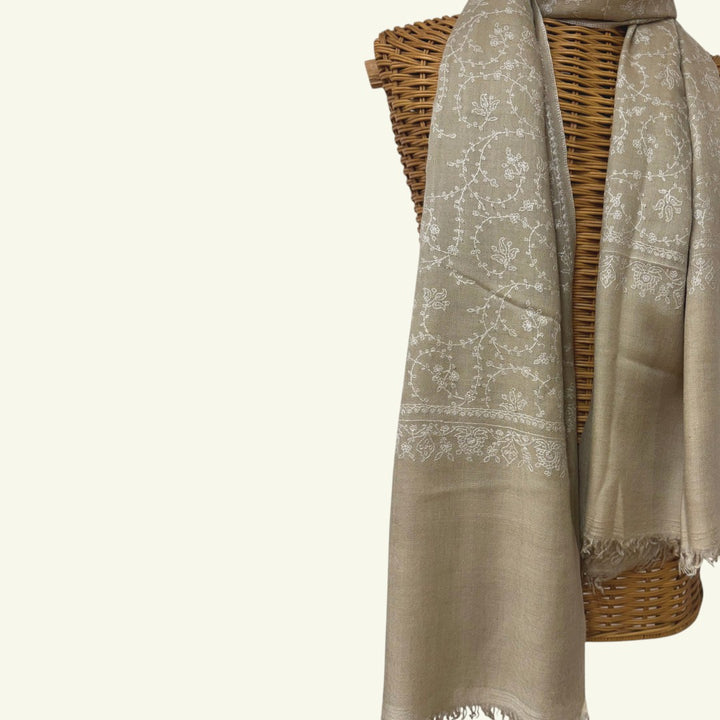 A light taupe-beige shawl with white floral embroidery and fringed edges, draped over a brown woven wicker basket against a plain off-white background.