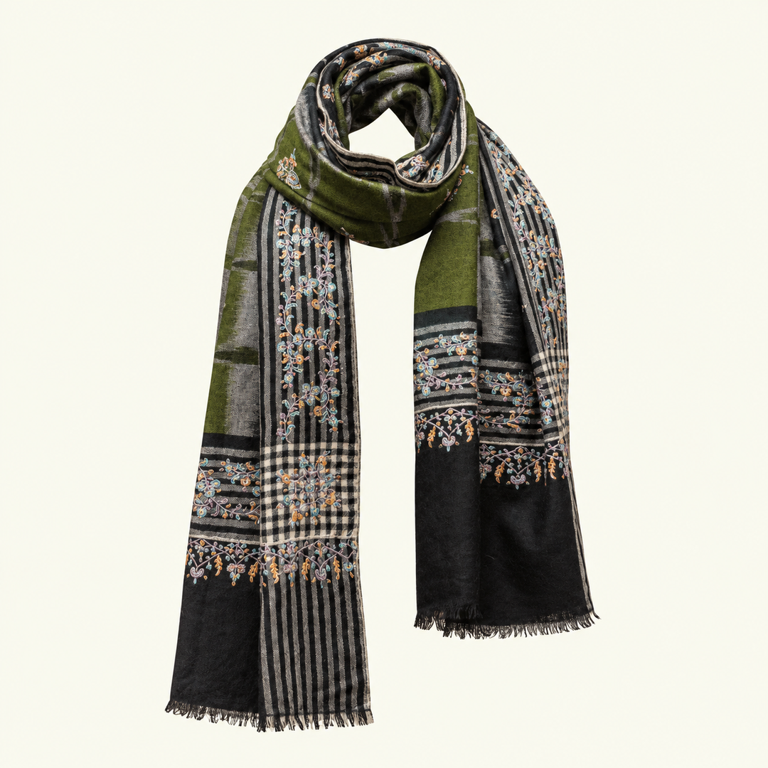 A long scarf with a complex patterned design featuring olive green, black, beige, and gray blocks, stripes, and floral embroidery with small orange and blue accents, finished with fringe on a light cream background.