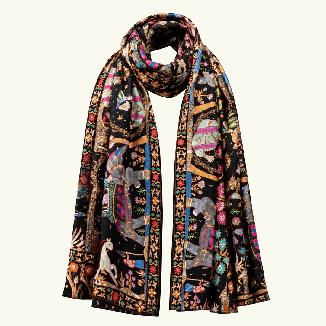 A long pashmina scarf with an intricate, colorful tapestry-style print featuring elephants, figures, and floral motifs on a black background, draped against a solid off-white background.