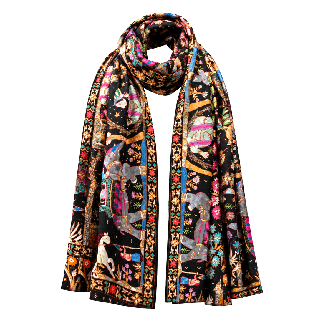 A long pashmina scarf with an intricate, colorful tapestry-style print featuring elephants, figures, and floral motifs on a black background, draped against a solid off-white background.