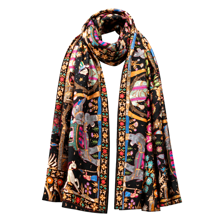 A long pashmina scarf with an intricate, colorful tapestry-style print featuring elephants, figures, and floral motifs on a black background, draped against a solid off-white background.